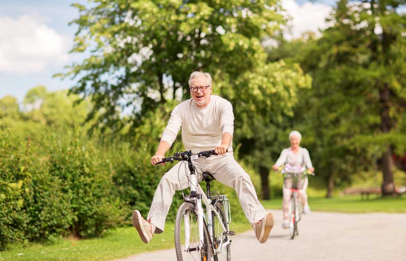 Older man riding a bicycle with his legs outstretched, smiling, with an older woman cycling behind him on a sunny path in a park with greenery and trees.