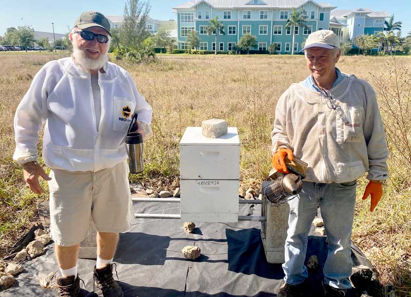 Two beekeepers wearing protective gear stand beside a white beehive in a grassy field, holding smokers. Buildings are visible in the background.
